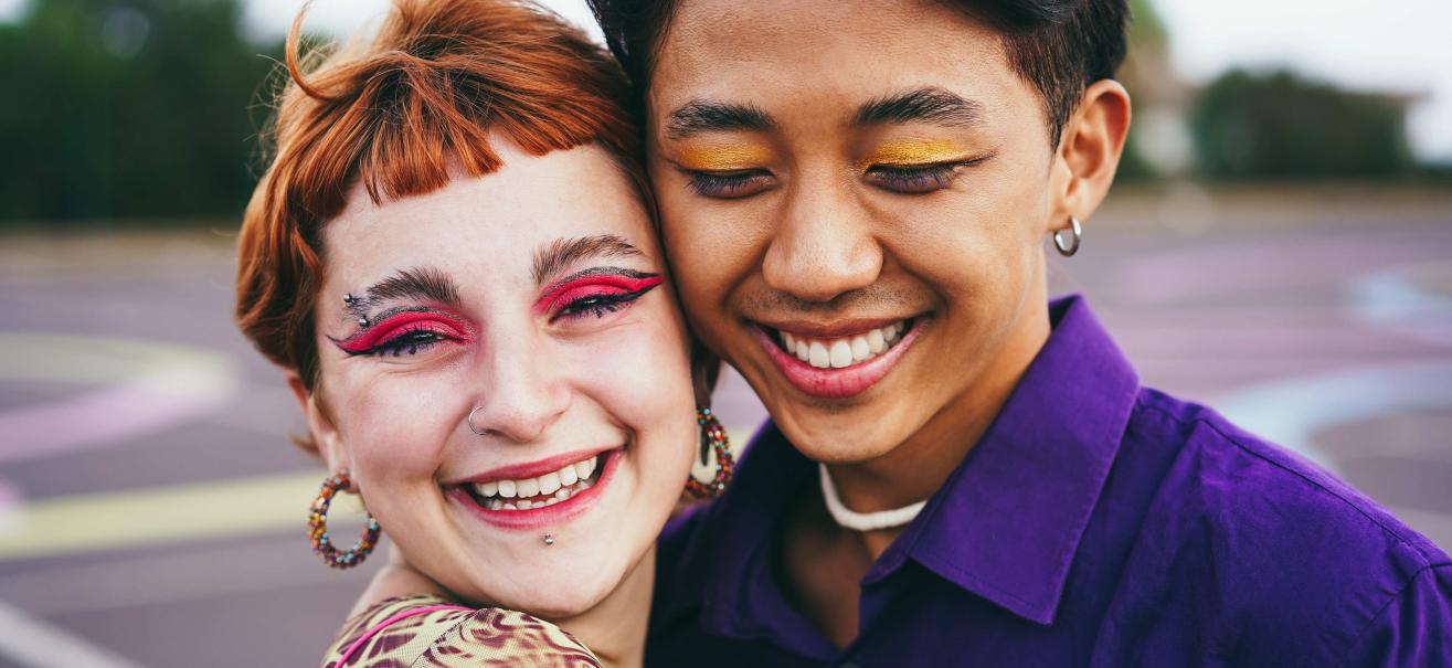 Two people between the ages of 17-25 stand temple to temple smiling at the camera. One person has red hair and pink eye shadow, and the other has black hair with a blue collared shirt. 