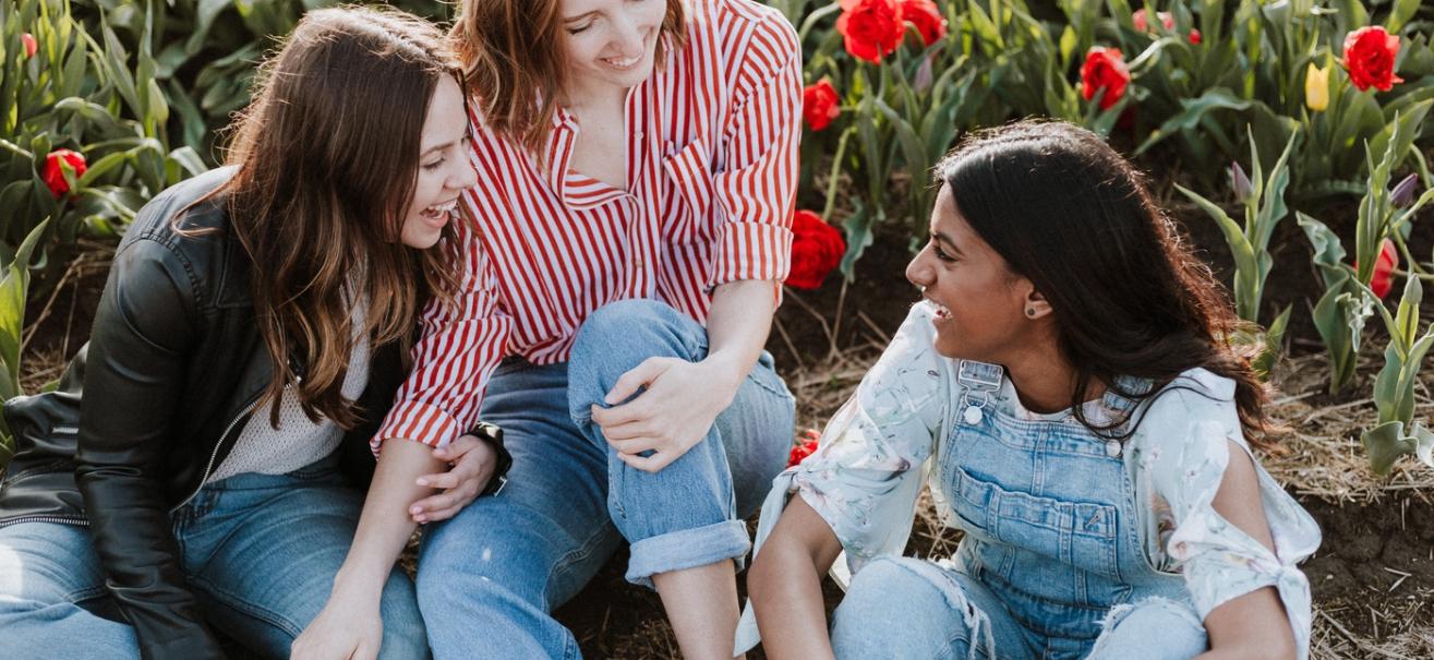 Three young people assigned female at birth laugh together while sitting outside a garden in the daytime