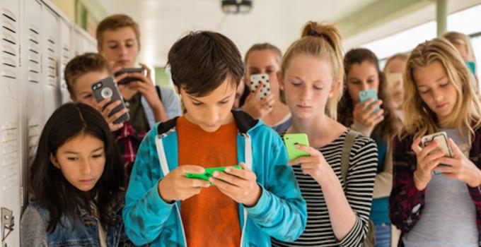 middle schoolers stand in a school hallway by lockers looking down at their phones