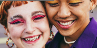 A white female with red hair, dark brown eyebrows, and bright pink eyeshadow smiles at the camera. She is touching cheeks with a brown male who is wearing yellow eyeshadow. His eyes are closed, and he is smiling at the camera.