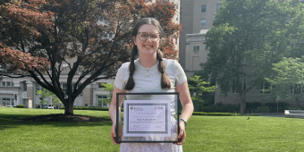 A white female with long brown hair smiling at the camera and holding a frame with a certificate in it. She is standing in front of a tree with red leaves, green grass, and a short stone wall.