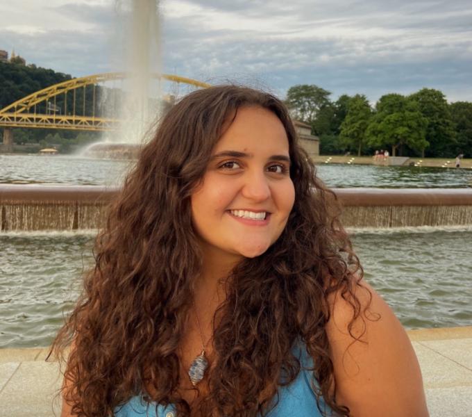 Dominique is a twenty-something cis woman with long curly brown hair, standing in front of a fountain outside. 