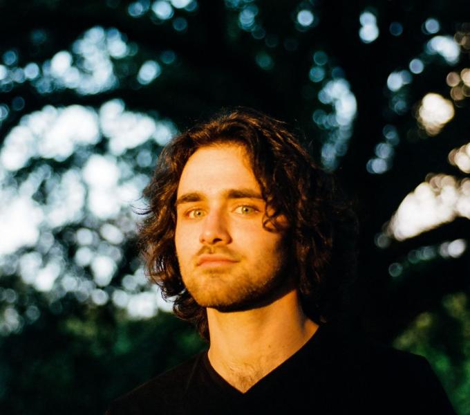 A white male with chin-length black hair looking at the camera with golden hour light shining on his face with a tree behind him.
