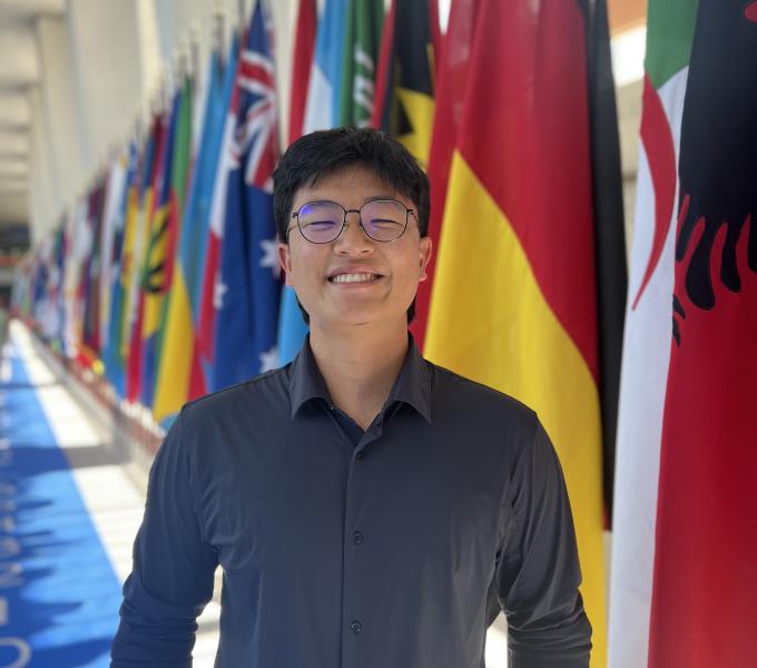 A young, Asian man wearing a gray shirt and glasses standing in front of a collection of flags