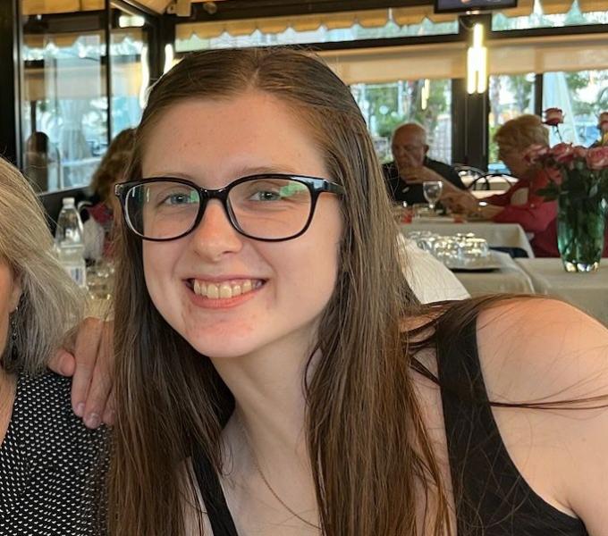 A white girl with dark brown glasses and long brown hair smiling at the camera.
